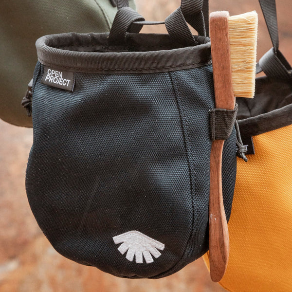 Black chalk bag with a logo, attached to a climbing harness, with a brush on top against a blurred natural background.